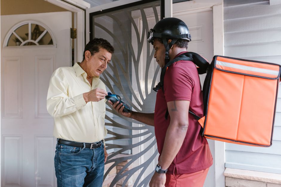A male customer holding a mobile phone receives a flower delivery from a delivery person wearing a maroon uniform, helmet, and a large orange insulated delivery bag on their back, outside a residentia