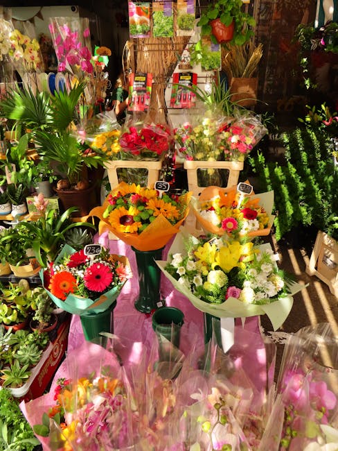 A colorful display of fresh flower bouquets arranged in various vases and wrapping papers on a pink table within a florist shop. The bouquets include bright yellow sunflowers, pink and white roses, re