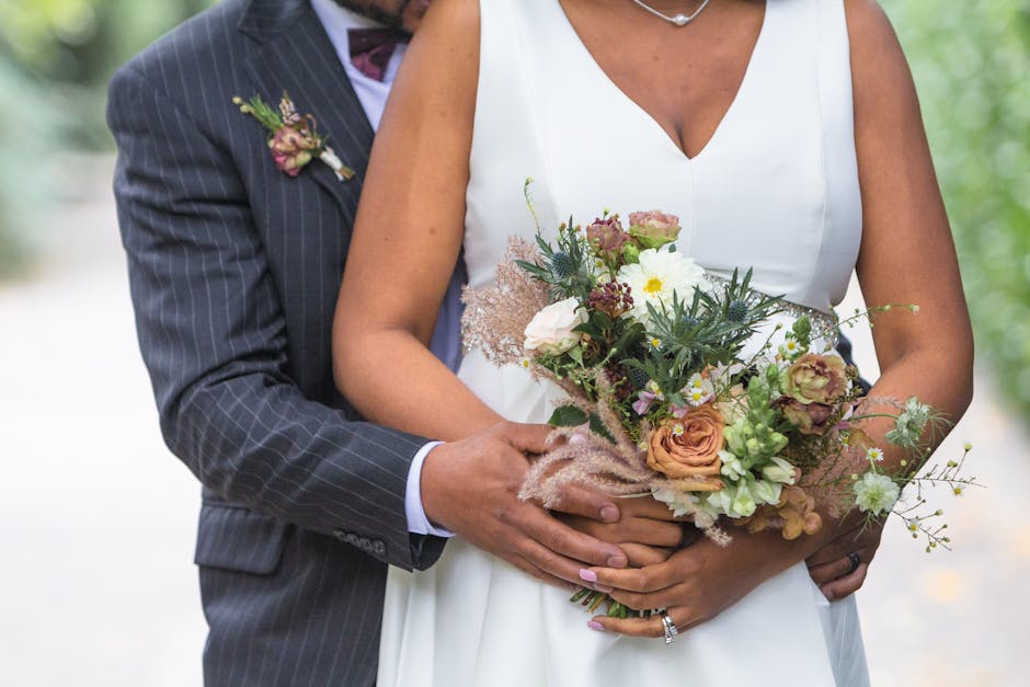 Close-up of a bride and groom holding a floral bouquet during a wedding ceremony near Haggerston Park. The flower arrangement features soft peach and white roses, delicate daisies, and various greener