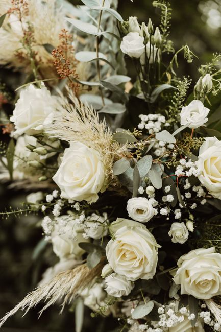 A floral arrangement featuring large, creamy white roses with full, layered petals, complemented by smaller white blooms and delicate sprigs of baby's breath. The bouquet includes silvery-green eucaly